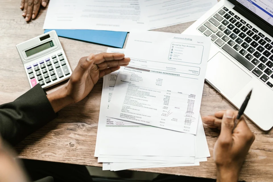 A person holding printed financial documents beside a calculator and laptop.