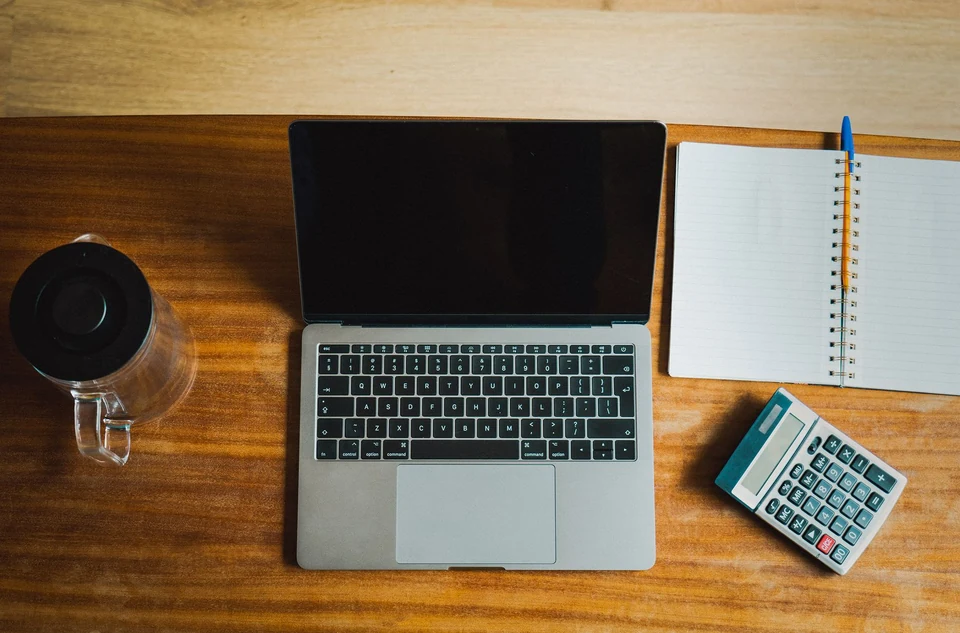 An overhead view of a laptop, calculator and notebook on a wooden desk.